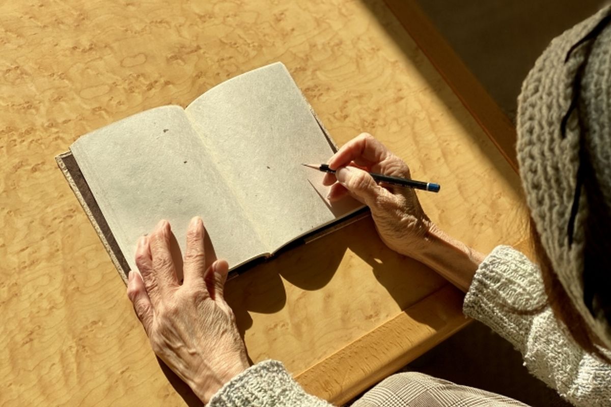机に向かってノートを広げ、何かを書き込んでいる老婆を斜め上から撮影した様子。エンディングノートや生前整理を象徴するイメージ。